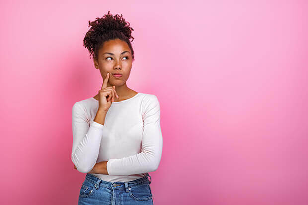 A young Black woman in front of a pink background furrowing her brow in concentration as if thinking hard about something