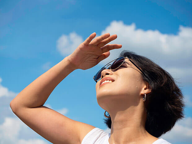 An East Asian woman shading her eyes and looking up toward a blue sky with clouds